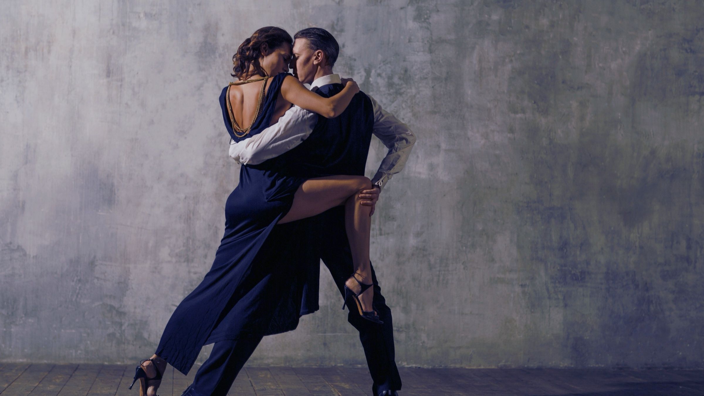 A couple in a dramatic leg-wrap dance pose against a grey concrete wall