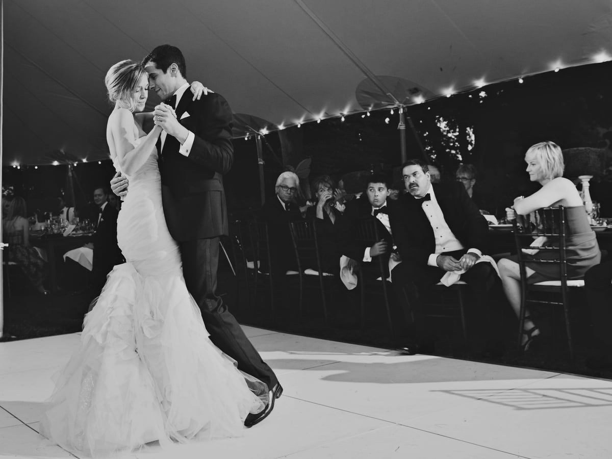 Black-and-white photo of a couple's first dance under a string-lit tent as guests watch